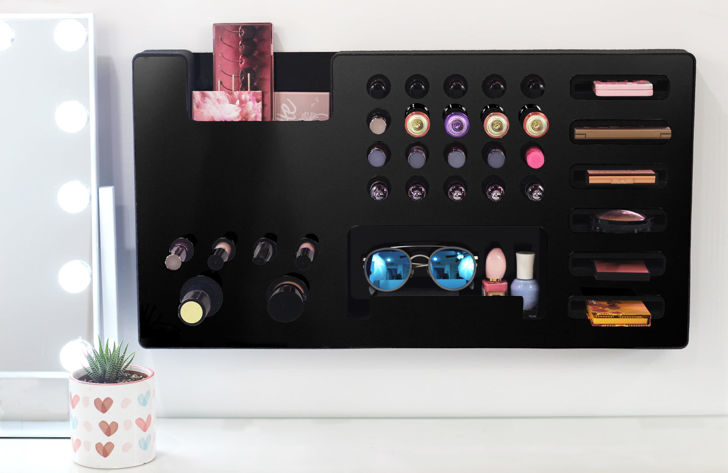 Black Ultra Complete Makeup Solution Organizer shown mounted on the wall holding lipsticks, lip glosses, nail polishes, brushes, sunglasses and more by keeping items accessible and off of the counter. Shown next to a makeup vanity with samples of suggested items the organizer can hold.