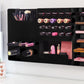 Black Ultra Complete Makeup Solution Organizer shown mounted on the wall holding lipsticks, lip glosses, nail polishes, brushes, sunglasses and more by keeping items accessible and off of the counter. Shown next to a makeup vanity with samples of suggested items the organizer can hold. Tilted to the left to show depth.