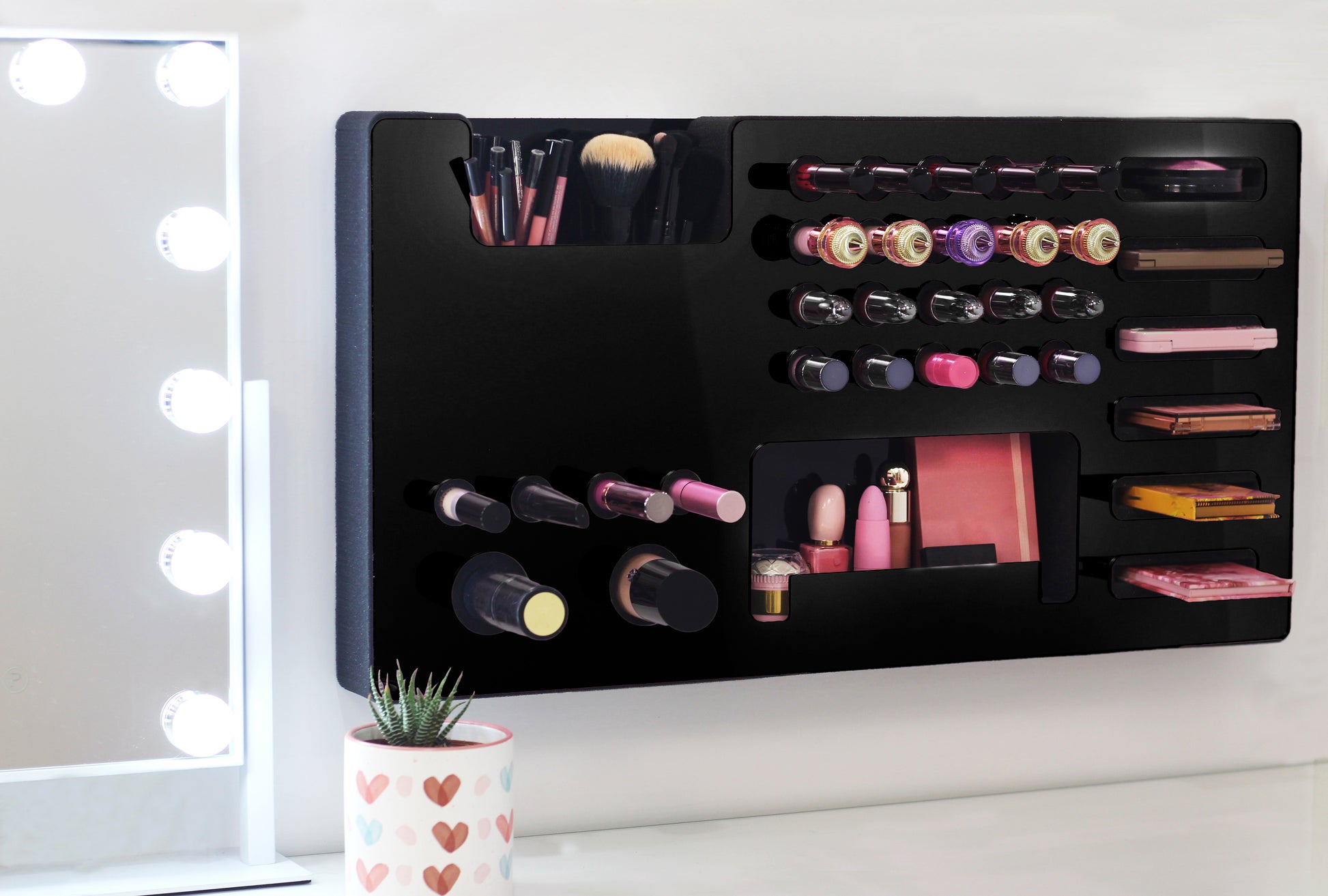 Black Ultra Complete Makeup Solution Organizer shown mounted on the wall holding lipsticks, lip glosses, nail polishes, brushes, sunglasses and more by keeping items accessible and off of the counter. Shown next to a makeup vanity with samples of suggested items the organizer can hold. Tilted to the right to show the wall mounted makeup organizer at a different angle while still on the wall.