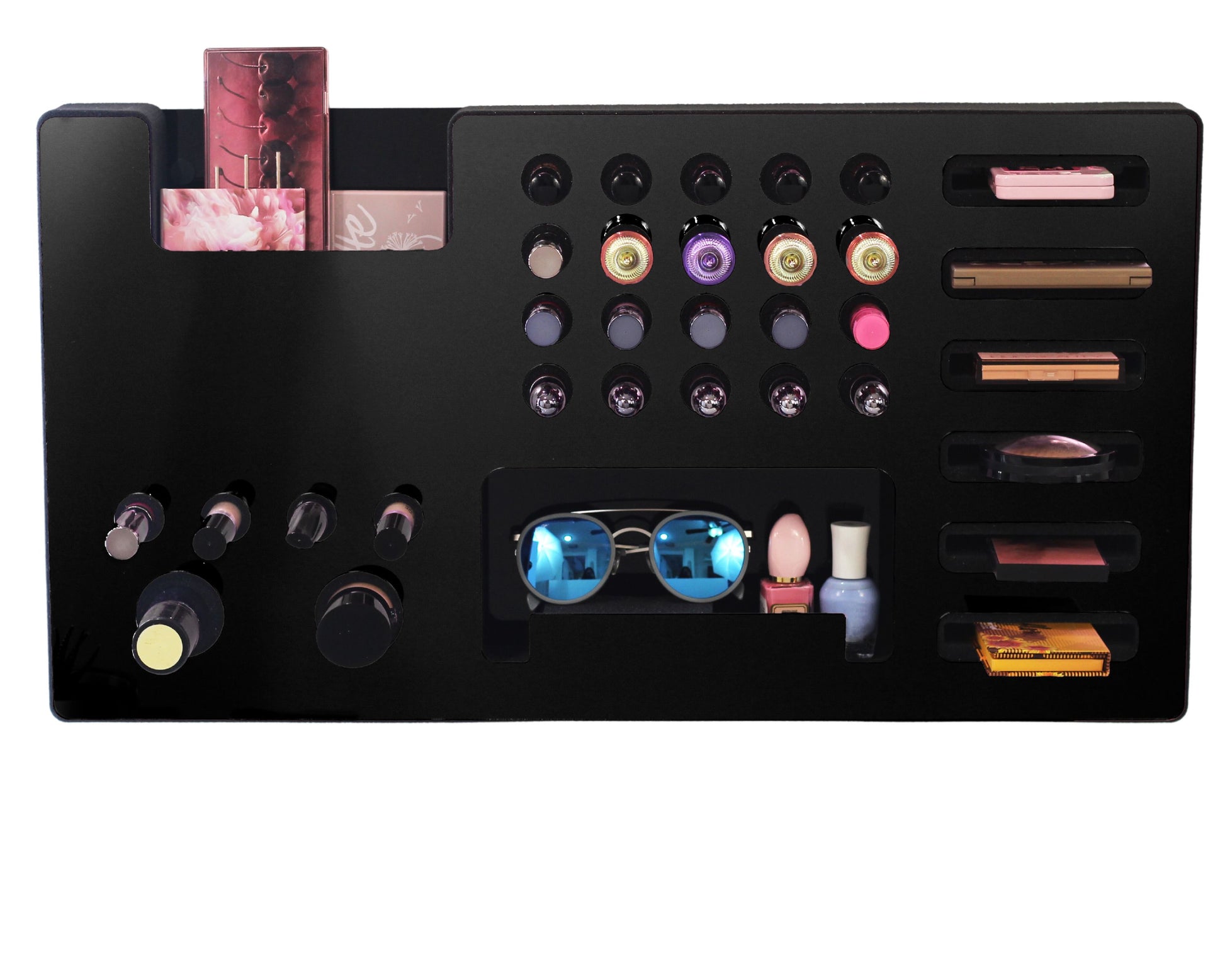 Black Ultra Complete Makeup Solution Organizer shown mounted on the wall holding lipsticks, lip glosses, nail polishes, brushes, sunglasses and more by keeping items accessible and off of the counter. Shown next to a makeup vanity with samples of suggested items the organizer can hold. Shown with a white background.