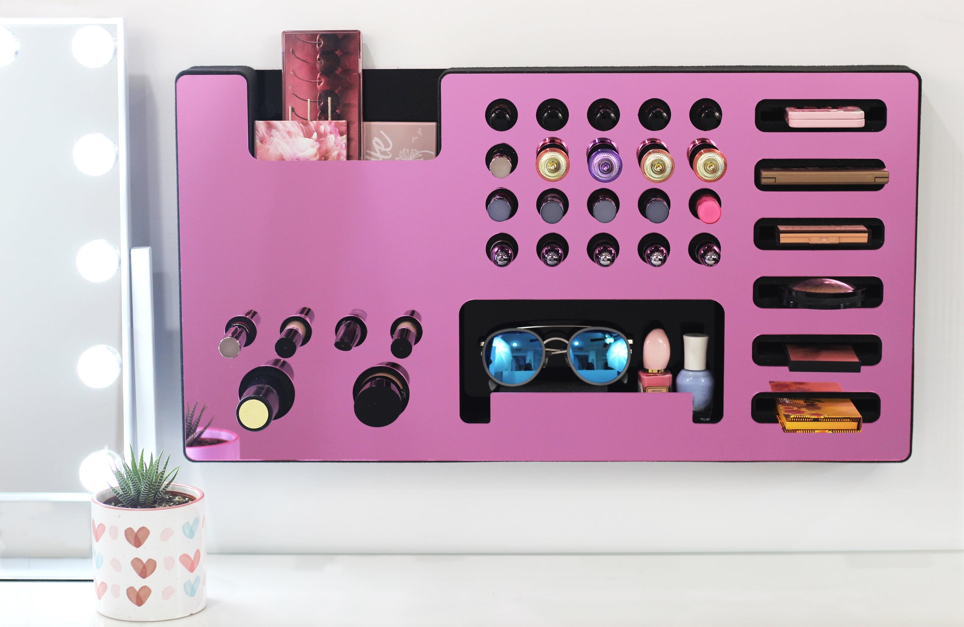Mirror Pink Ultra Complete Makeup Solution Organizer shown mounted on the wall holding lipsticks, lip glosses, nail polishes, brushes, sunglasses and more by keeping items accessible and off of the counter. Shown next to a makeup vanity with samples of suggested items the organizer can hold.