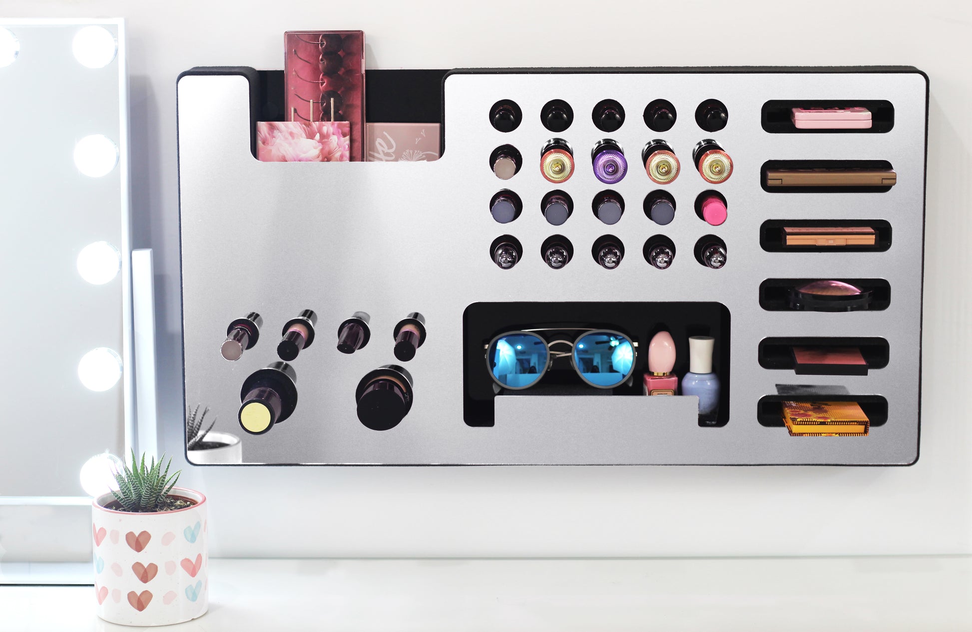 Mirror Silver Ultra Complete Makeup Solution Organizer shown mounted on the wall holding lipsticks, lip glosses, nail polishes, brushes, sunglasses and more by keeping items accessible and off of the counter. Shown next to a makeup vanity with samples of suggested items the organizer can hold.