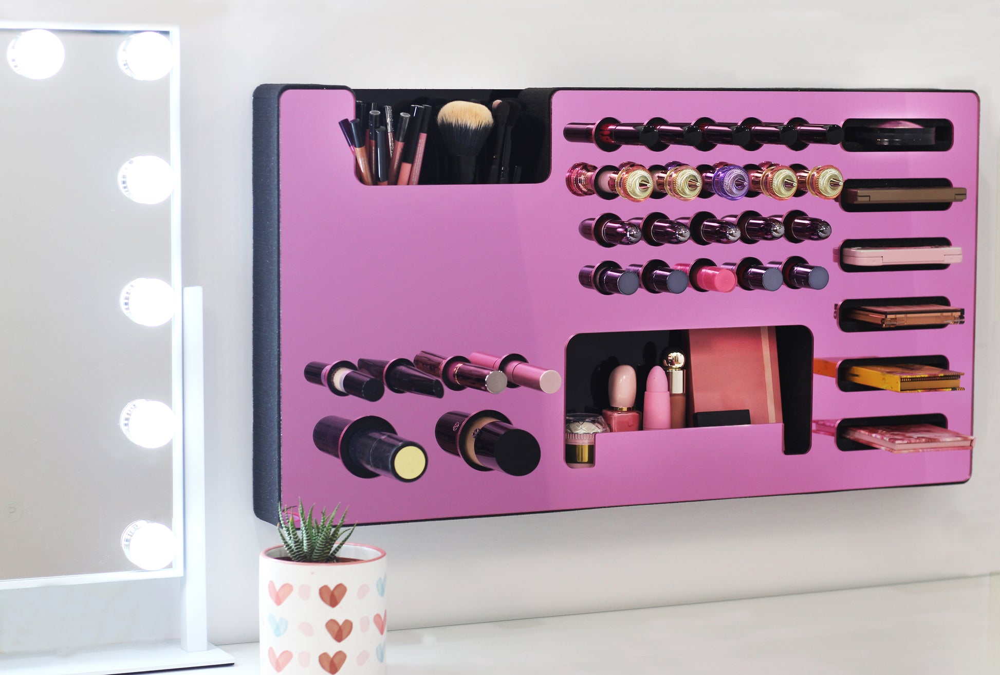 Mirror Pink Ultra Complete Makeup Solution Organizer shown mounted on the wall holding lipsticks, lip glosses, nail polishes, brushes, sunglasses and more by keeping items accessible and off of the counter. Shown next to a makeup vanity with samples of suggested items the organizer can hold. Tilted to the right to show the wall mounted makeup organizer at a different angle while still on the wall.