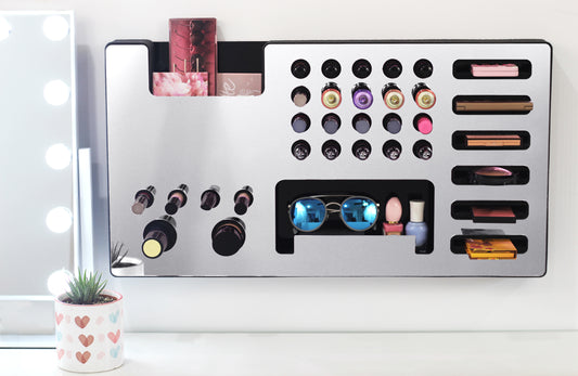 Mirror Silver Ultra Complete Makeup Solution Organizer shown mounted on the wall holding lipsticks, lip glosses, nail polishes, brushes, sunglasses and more by keeping items accessible and off of the counter. Shown next to a makeup vanity with samples of suggested items the organizer can hold.