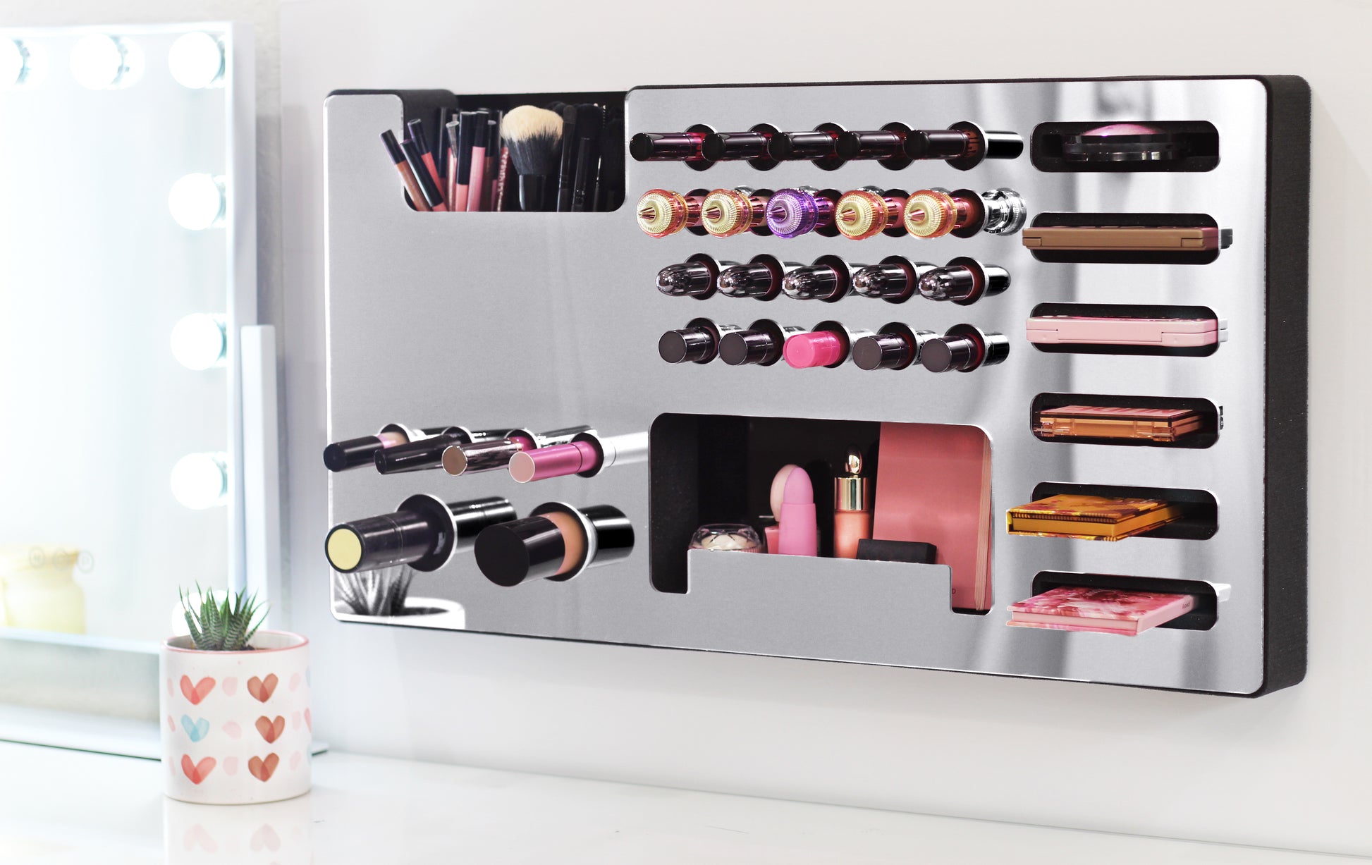 Mirror Silver Ultra Complete Makeup Solution Organizer shown mounted on the wall holding lipsticks, lip glosses, nail polishes, brushes, sunglasses and more by keeping items accessible and off of the counter. Shown next to a makeup vanity with samples of suggested items the organizer can hold. Tilted to the left to show depth.