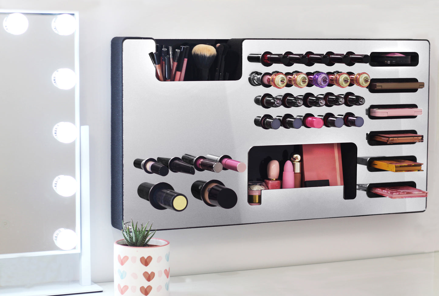 Mirror Silver Ultra Complete Makeup Solution Organizer shown mounted on the wall holding lipsticks, lip glosses, nail polishes, brushes, sunglasses and more by keeping items accessible and off of the counter. Shown next to a makeup vanity with samples of suggested items the organizer can hold. Tilted to the right to show the wall mounted makeup organizer at a different angle while still on the wall.