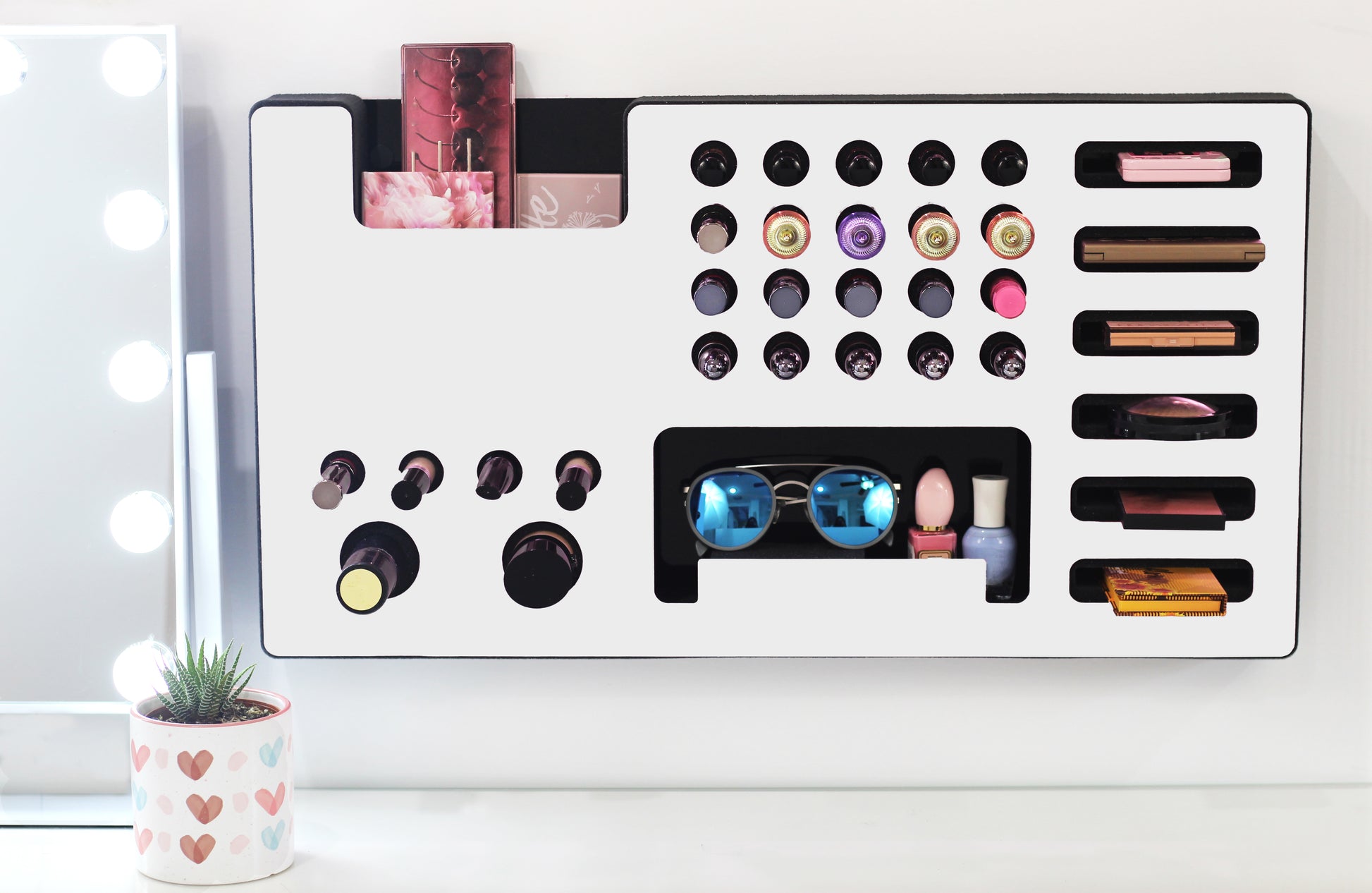 White Ultra Complete Makeup Solution Organizer shown mounted on the wall holding lipsticks, lip glosses, nail polishes, brushes, sunglasses and more by keeping items accessible and off of the counter. Shown next to a makeup vanity with samples of suggested items the organizer can hold.