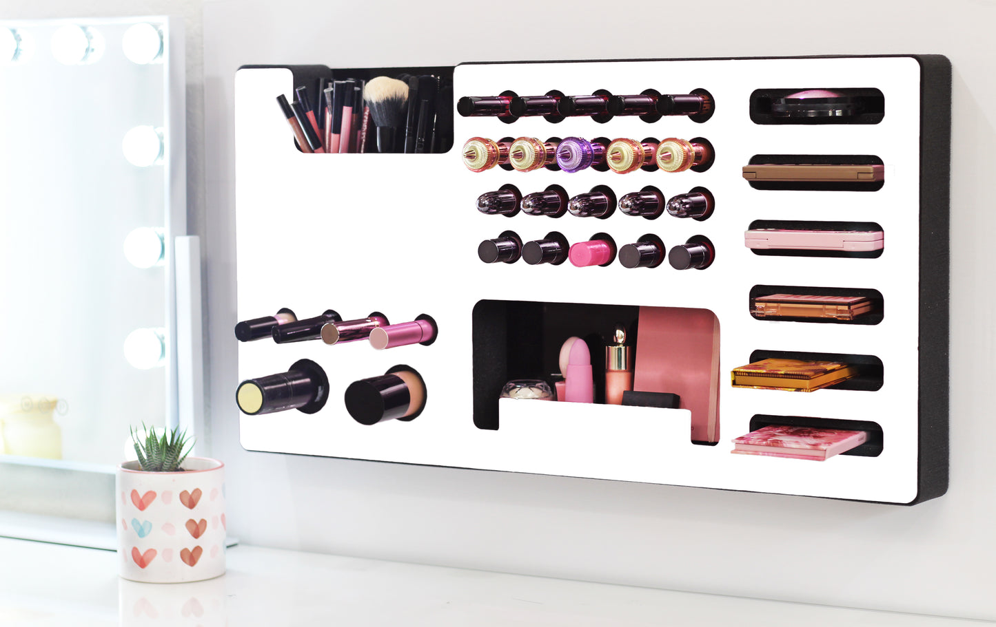 White Ultra Complete Makeup Solution Organizer shown mounted on the wall holding lipsticks, lip glosses, nail polishes, brushes, sunglasses and more by keeping items accessible and off of the counter. Shown next to a makeup vanity with samples of suggested items the organizer can hold. Tilted to the left to show depth.